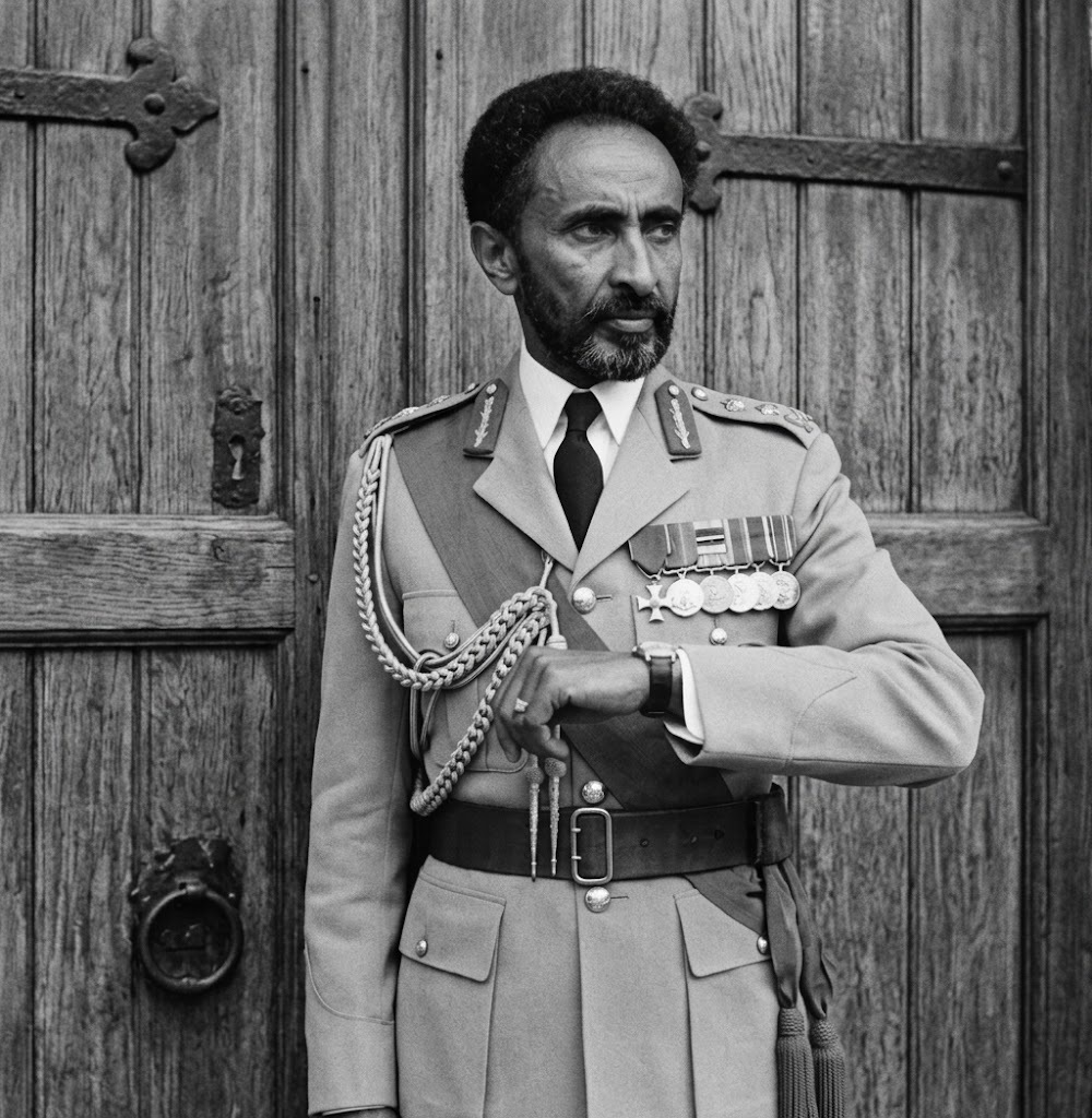 Haile Selassie I in military uniform, checking his wristwatch outside an audience chamber